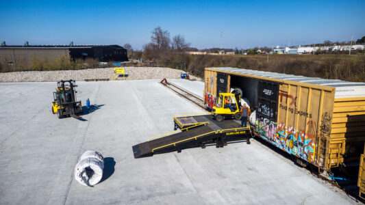 A flat bed truck with a ramp is pushed against a box car. Next to the ramp is a wrapped steel wire that has just been unloaded from the boxcar. 