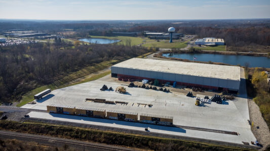 A drone shot of King Steel's logistics warehouse, right next to rail siding. On the rail siding are four box cars. 